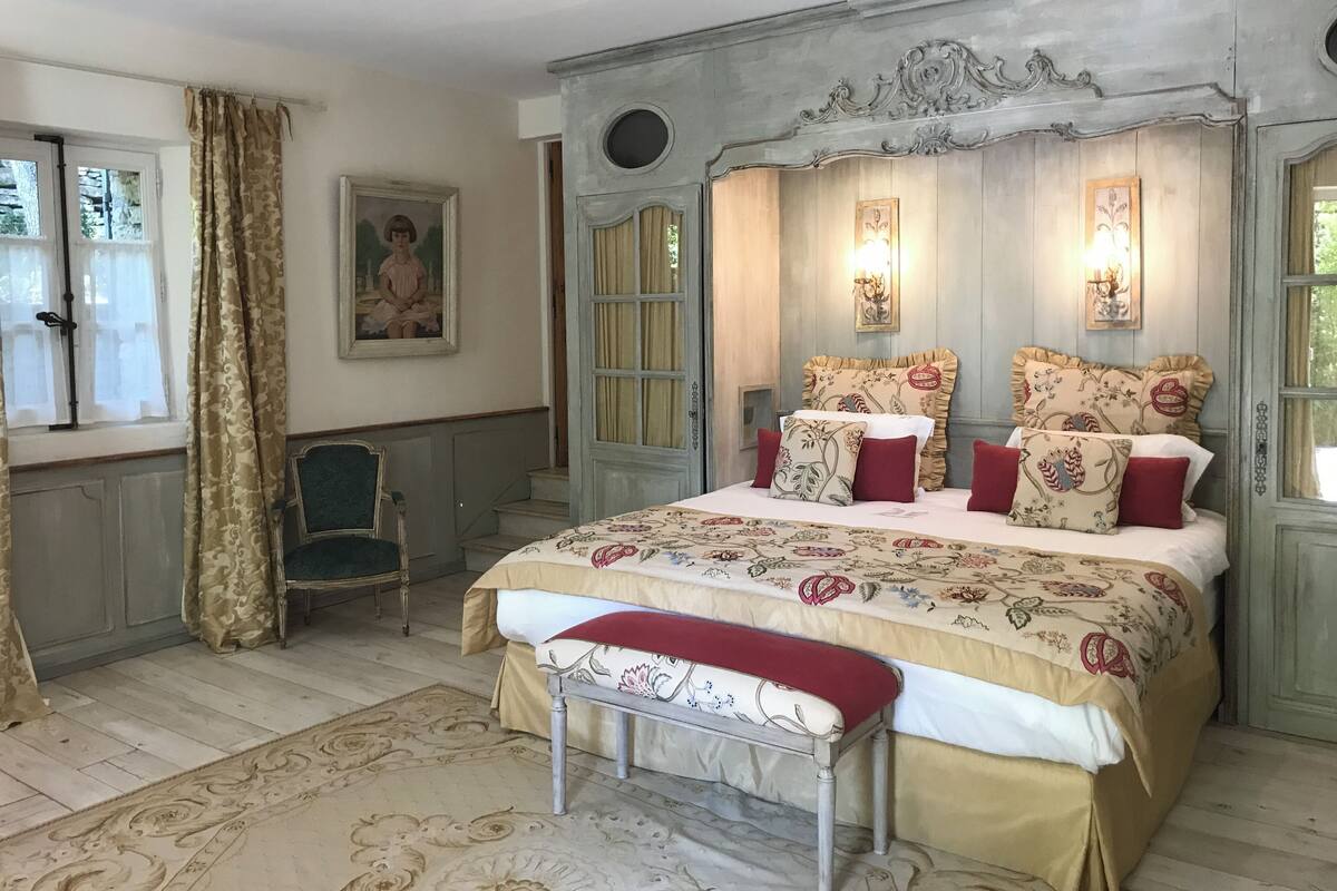 A bedroom with a large bed, a chair, a bench, and a window with curtains.