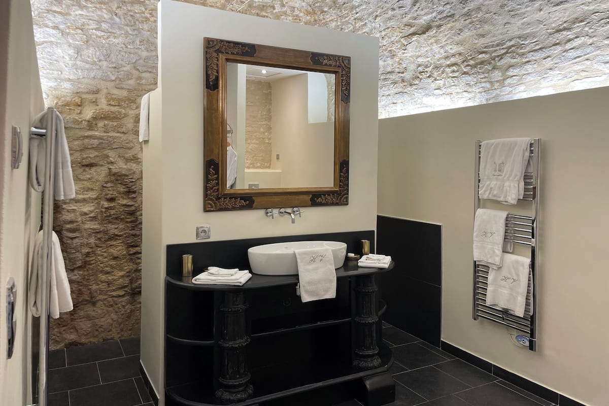 A bathroom with a stone wall, a large mirror, a black vanity, a white sink, and a towel rack.