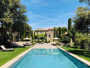 A swimming pool with lounge chairs and umbrellas, surrounded by well-maintained greenery and a stone building in the background. - Villa Hautvallon (Gordes)