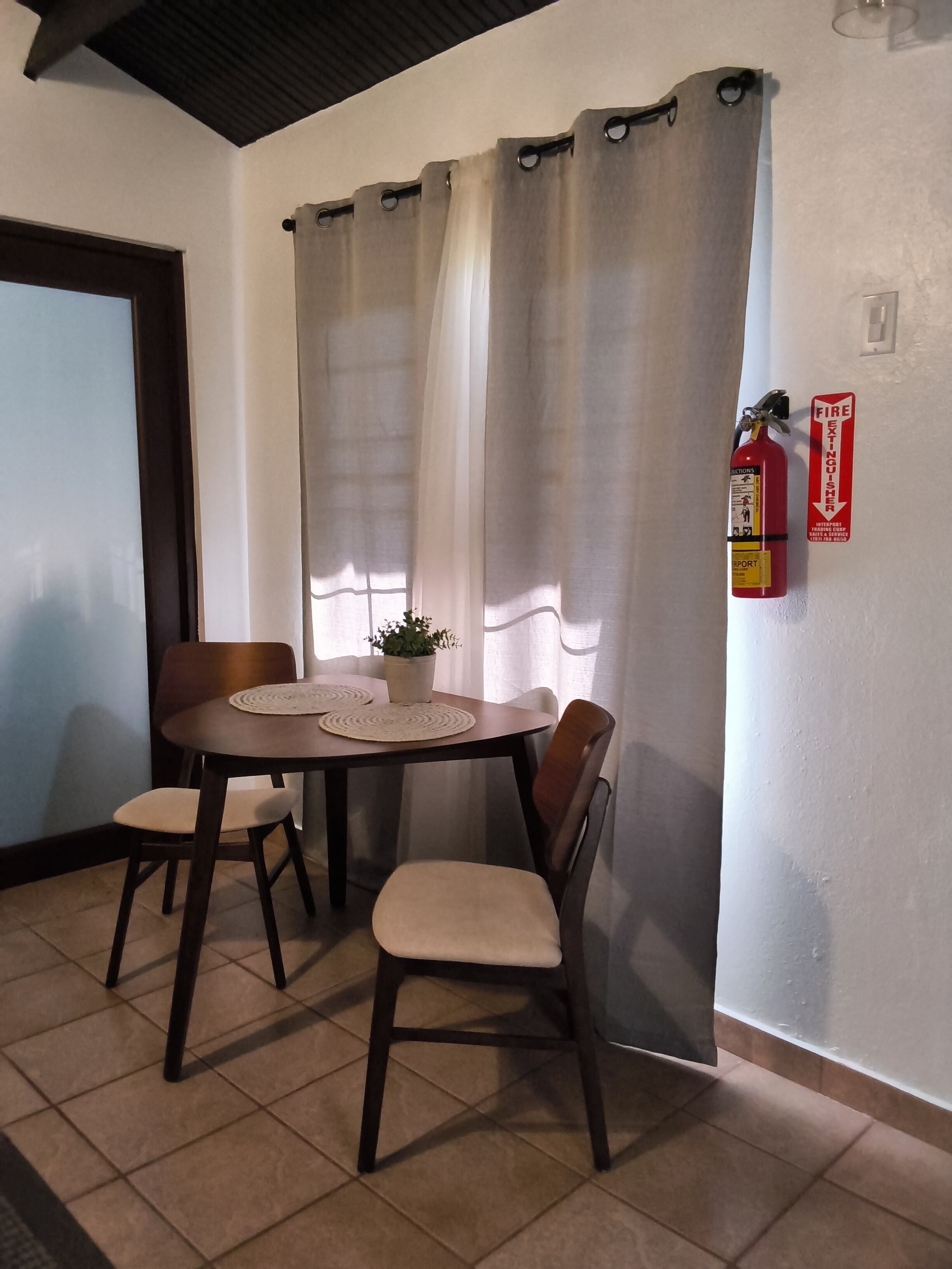 Ein Essbereich mit Tisch, Stühlen, einer Topfpflanze und einem an der Wand befestigten Feuerlöscher.