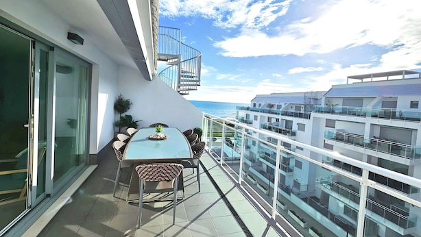 Un balcon avec une table en verre et des chaises en osier, offrant une vue sur un immeuble moderne et la mer.
