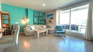 Un salon aux murs de couleur turquoise, avec un canapé, une table basse et un fauteuil. On y trouve des étagères ornées d’objets décoratifs et une grande fenêtre offrant une belle vue.