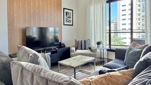 A modern living room with a large flat-screen TV, a sofa, a coffee table, and a view of tall buildings through a large window. - Amazing apartment near Blue Mall with pool in Regatta Living YellowKey (Santo Domingo)