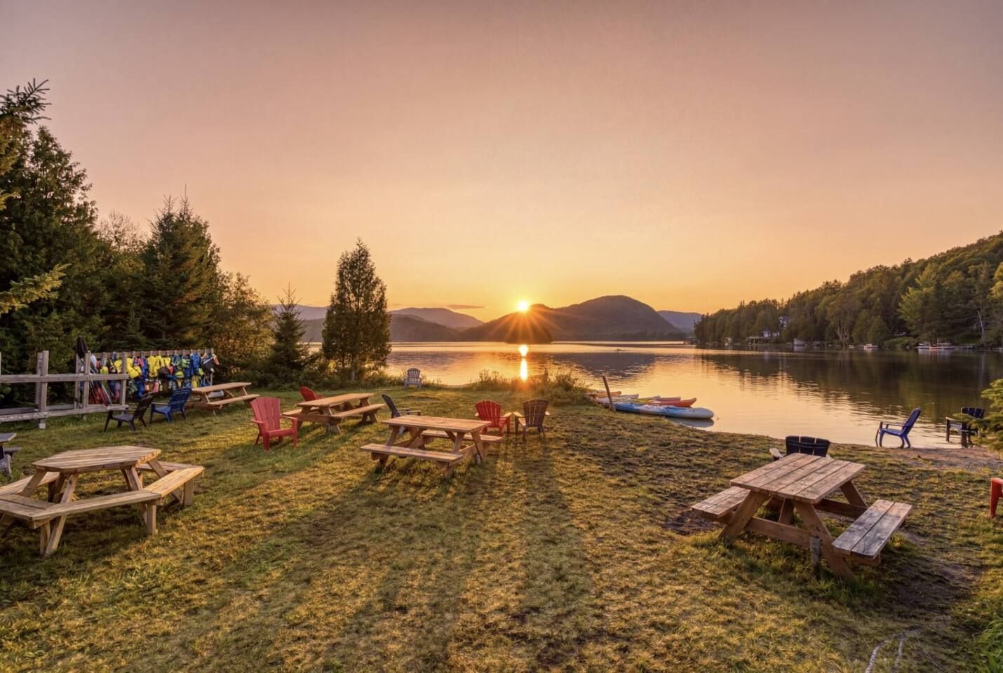 Un espace de pique-nique au bord du lac, avec des bancs et des chaises en bois, un canoë et une rangée de fauteuils Adirondack.