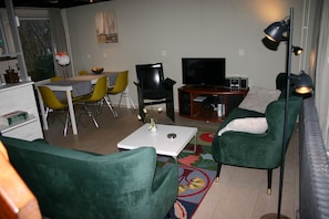 Ein Wohnzimmer mit grünen Sesseln, einem weißen Tisch, einem Fernseher und einem kleinen Schrank.