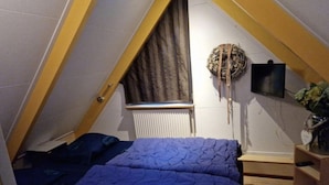 Une petite chambre mansardée de forme triangulaire, avec un plafond en pente, un radiateur, un téléviseur fixé au mur et un lit recouvert d’une couverture bleue.