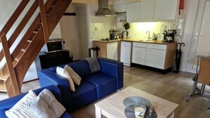 Un espace de vie compact comprenant un canapé bleu, une petite table, un coin cuisine et un escalier en bois.