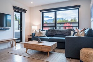 A modern living room with a sofa, coffee table, and a TV. A large window offers a view of mountains and trees. - Bluebird Daze (Revelstoke)