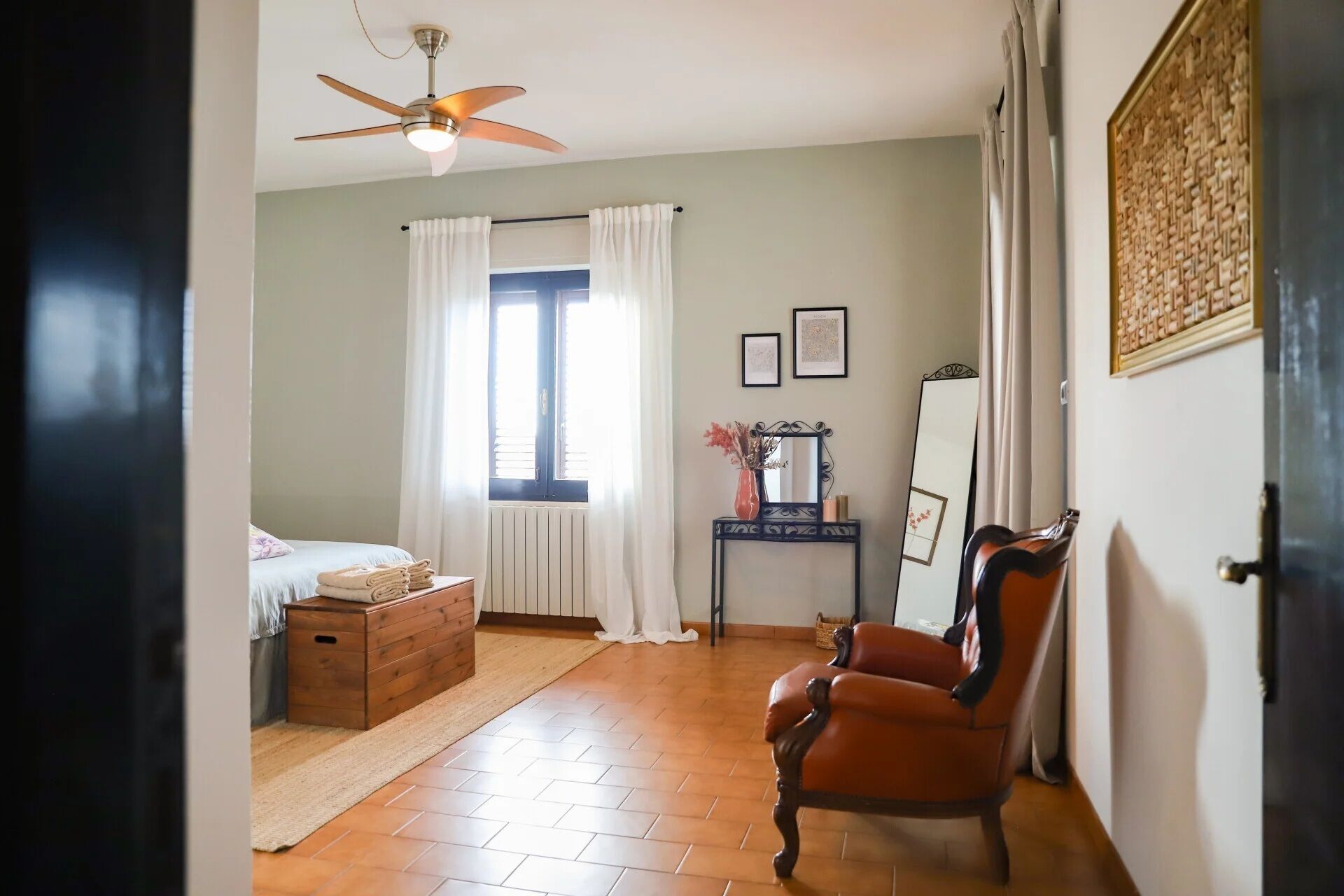 A bedroom with a bed, a wooden chest, a chair, a desk with a mirror, and a ceiling fan.