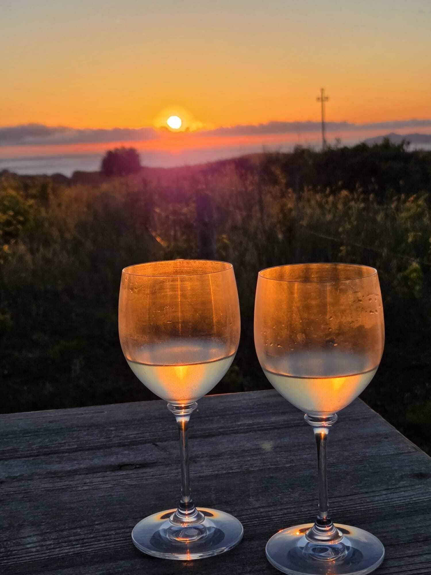 Dos copas de vino sobre una superficie de madera con un atardecer al fondo.