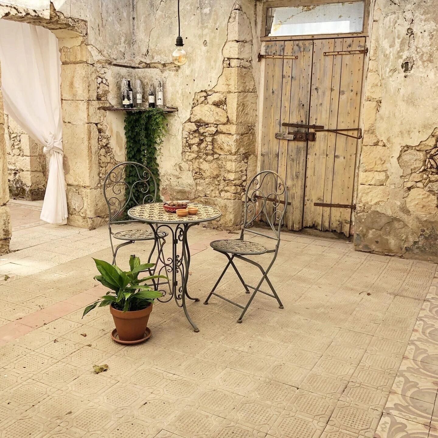 Une terrasse extérieure avec une table et des chaises en métal, un pot de fleurs et une porte en bois rustique.