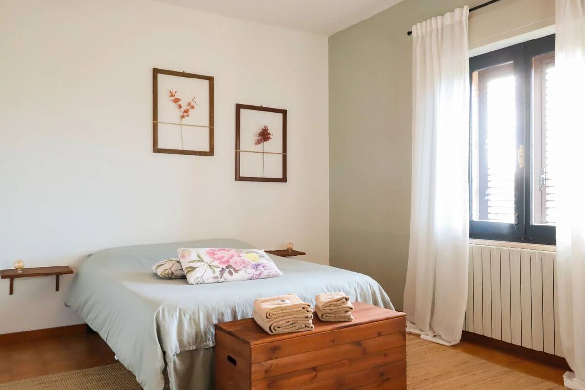 A bedroom with a bed, a wooden chest of drawers, a window with curtains, and two framed pictures on the wall.