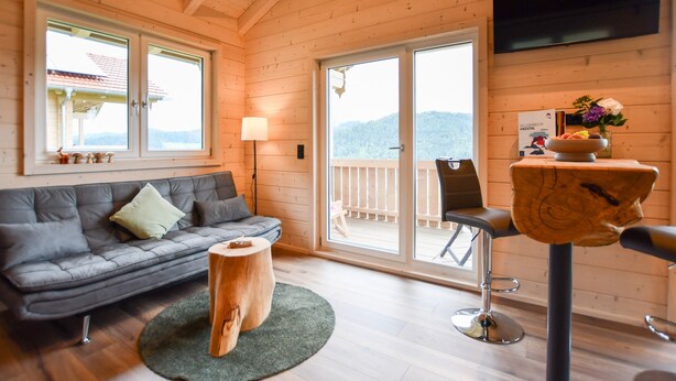 A wooden interior with a sofa, a round wooden table, a bar stool, a TV mounted on the wall, and a balcony with a view. - Holiday Home 'Panoramahisli 3' with Mountain View, Balcony and Air Conditioning (Fischerbach)