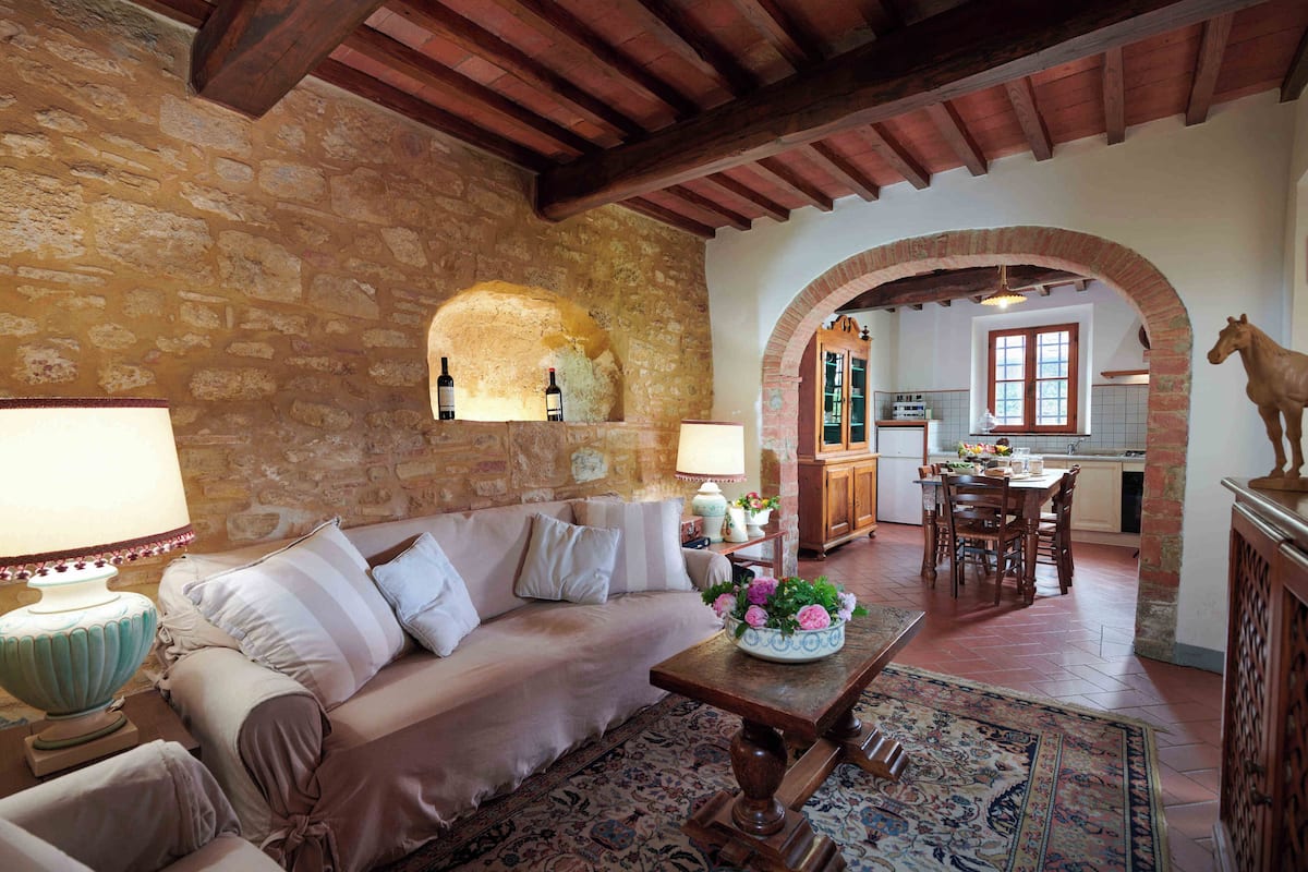 Un salon chaleureux avec un mur en pierre, un plafond en bois et un coin repas agrémenté d’une arche en briques.