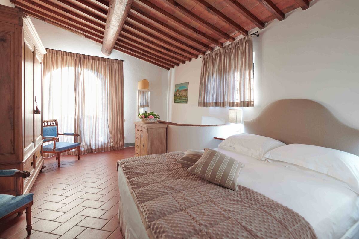 Une chambre avec un grand lit, une tête de lit en bois et un plafond en bois. On y trouve une chaise et une petite table sur laquelle est posé un vase de fleurs.