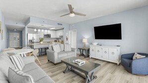 A modern living room with a grey sofa, a coffee table, and a blue armchair. A flat-screen TV is mounted on the wall, and a ceiling fan is visible. - Bright & Stylish Condo Near Everything! (Panama City Beach)
