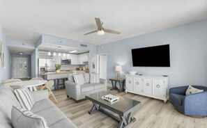 Ein modernes Wohnzimmer mit grauem Sofa, einem Couchtisch und einem blauen Sessel. Ein Flachbildfernseher ist an der Wand montiert, und ein Deckenventilator ist sichtbar.