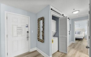 Ein Flur mit einer weißen Tür, einem Spiegel und einem Bett im angrenzenden Zimmer.