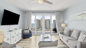 A modern living room with a flat-screen TV, a ceiling fan, a sofa, a coffee table, and a view of high-rise buildings through large windows. - Bright & Stylish Condo Near Everything! (Panama City Beach)
