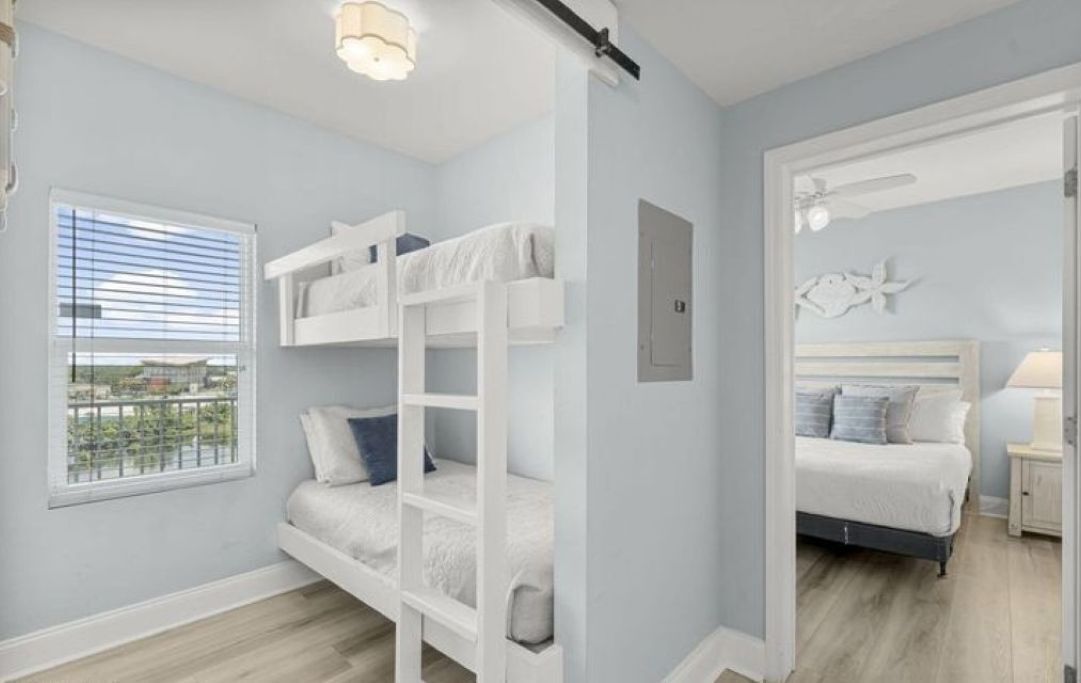 Une chambre avec des lits superposés, une fenêtre avec des stores, un lit avec une tête de lit et une table de chevet avec une lampe.
