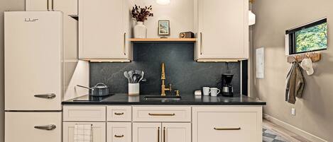 A kitchen with white cabinets, a black countertop, and a marble backsplash. There is a sink, a coffee maker, and a vase with dried flowers on the counter.
