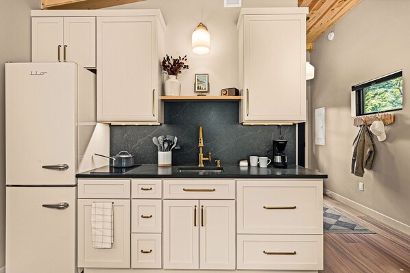 A kitchen with white cabinets, a black countertop, and a marble backsplash. There is a sink, a coffee maker, and a vase with dried flowers on the counter.