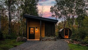 Une cabane moderne avec une façade en bois, une allée en gravier et une grande structure en forme de tonneau sur le côté droit.