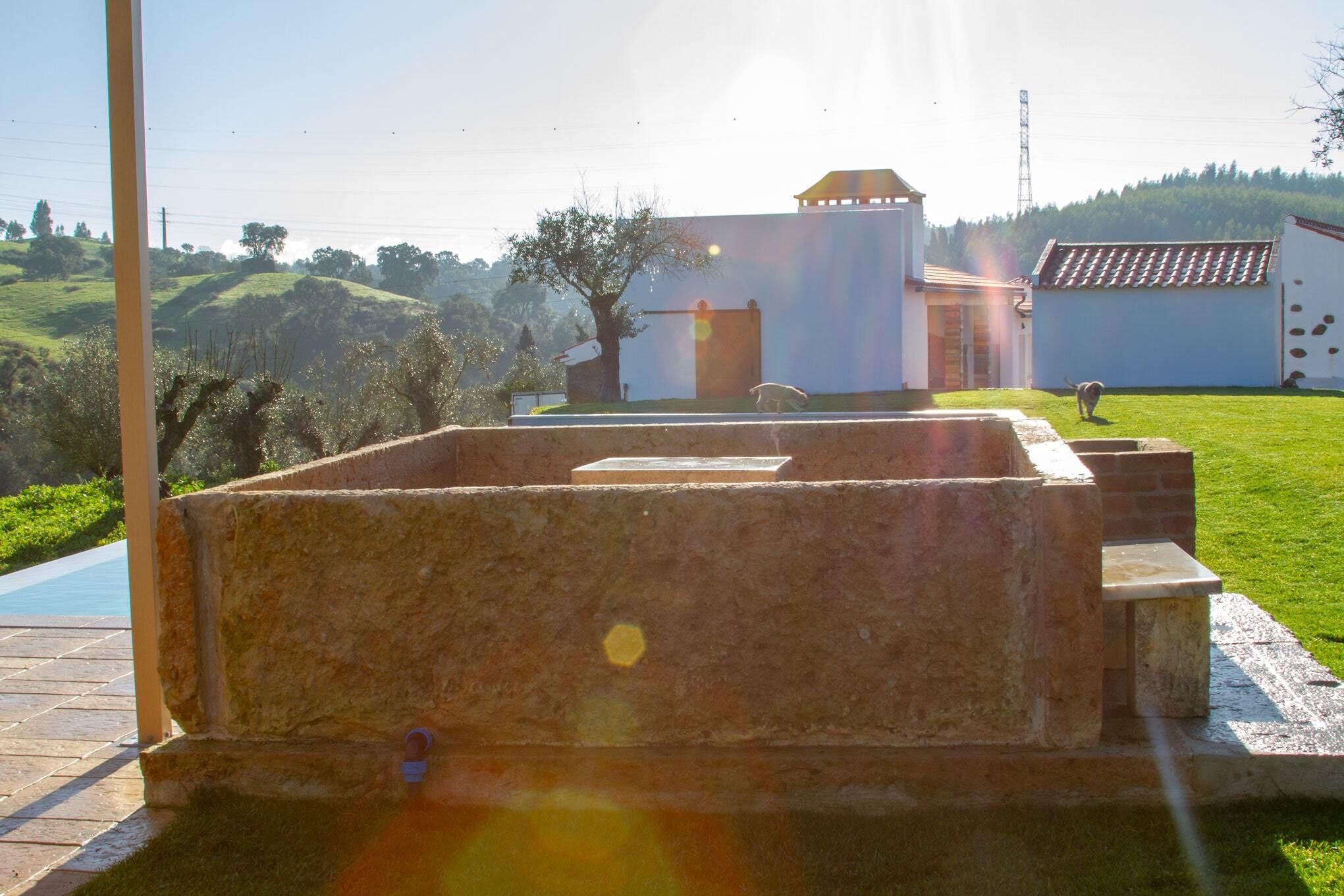 Una piscina con una estructura de piedra en primer plano, un edificio con techo de tejas al fondo y una zona ajardinada con árboles.