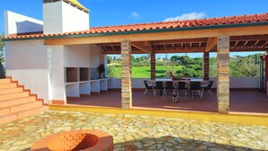 Une terrasse extérieure couverte, dotée d’un sol en pierre, d’un foyer en briques et d’un espace repas avec des chaises et une table.