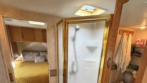 A compact RV bathroom with a shower enclosure, toilet, and a bed with a yellow bedspread. - Rv with 1 Bedrooms, and sofa bed Full Kitchen, Dining Area & Modern Bathroom” (Pikeville)