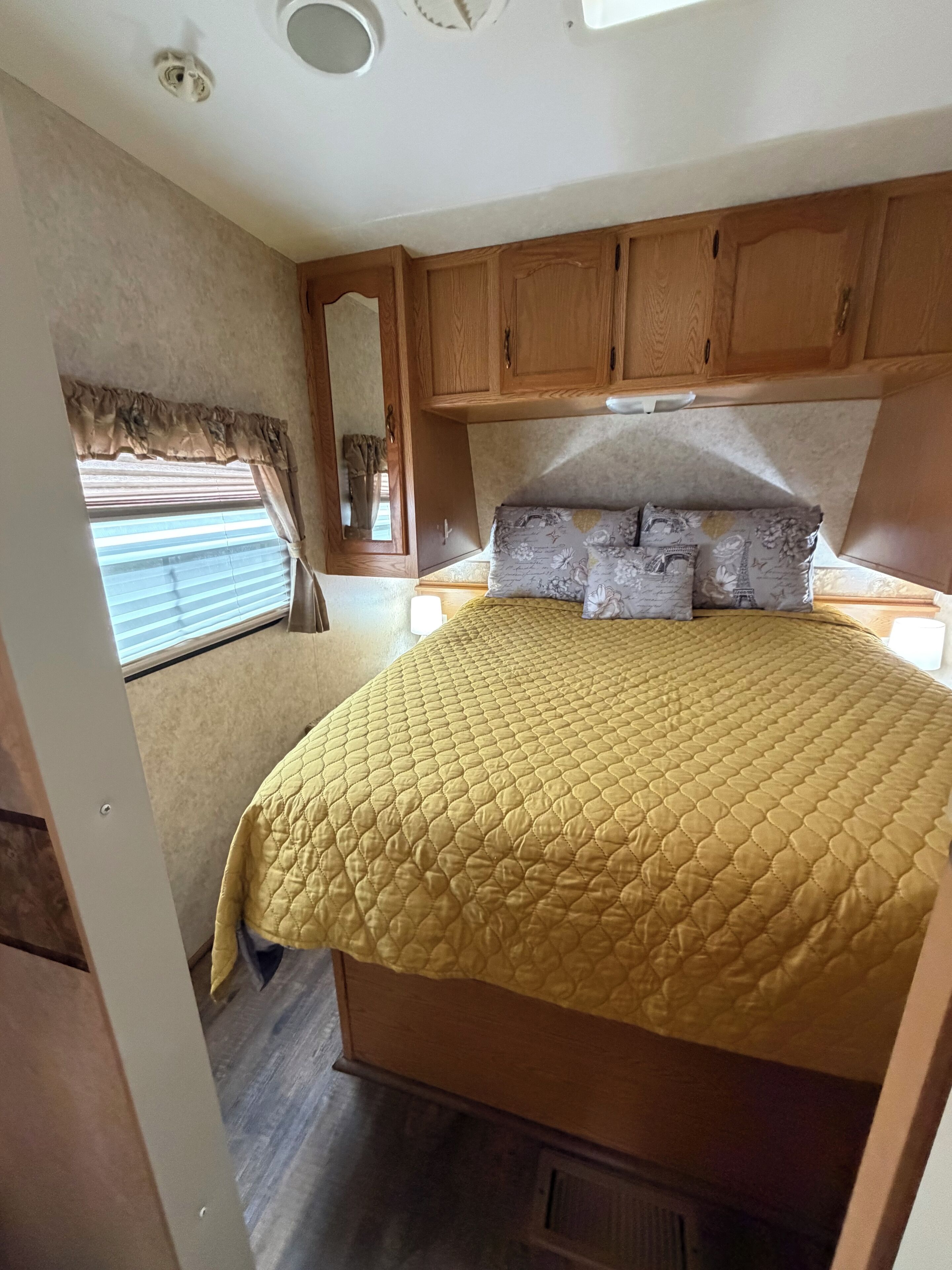 Un dormitorio de caravana compacto con cama, armarios de madera, ventana con persianas y rejilla de ventilación en el techo.