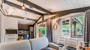 Un interior modern amb un sofà, taula de dinar i cadires, zona de cuina i grans finestres amb cortines.