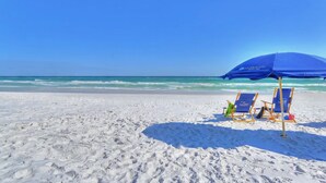 A beach with two blue lounge chairs under a blue umbrella, white sand, and the ocean in the background. - King's Castle | Luxury Beach Escape | Huge Private Pool/Spa | 6-Seater Golf Cart (Destin)