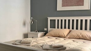 A neatly made bed with a white headboard and beige bedding, a bedside table with a lamp, and a framed picture on the wall. - Arte 36 (Bosa)