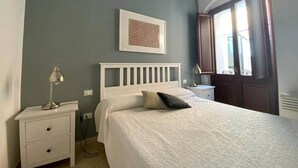 A bedroom with a bed, a nightstand, a lamp, a window with curtains, and a door. - Arte 36 (Bosa)