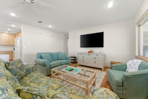 Ein Wohnzimmer mit Sofa, Couchtisch, Fernseher und einer Küchenzeile inklusive Schränke und Kühlschrank.