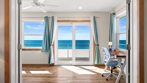 Ein Zimmer mit Holzboden, Deckenventilator, Schreibtisch mit Stuhl und Meerblick durch große Glasüren.
