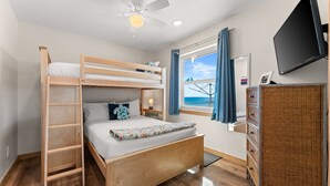 Un lit superposé avec une échelle en bois, un ventilateur de plafond, un téléviseur, une commode en osier et une fenêtre avec des rideaux.