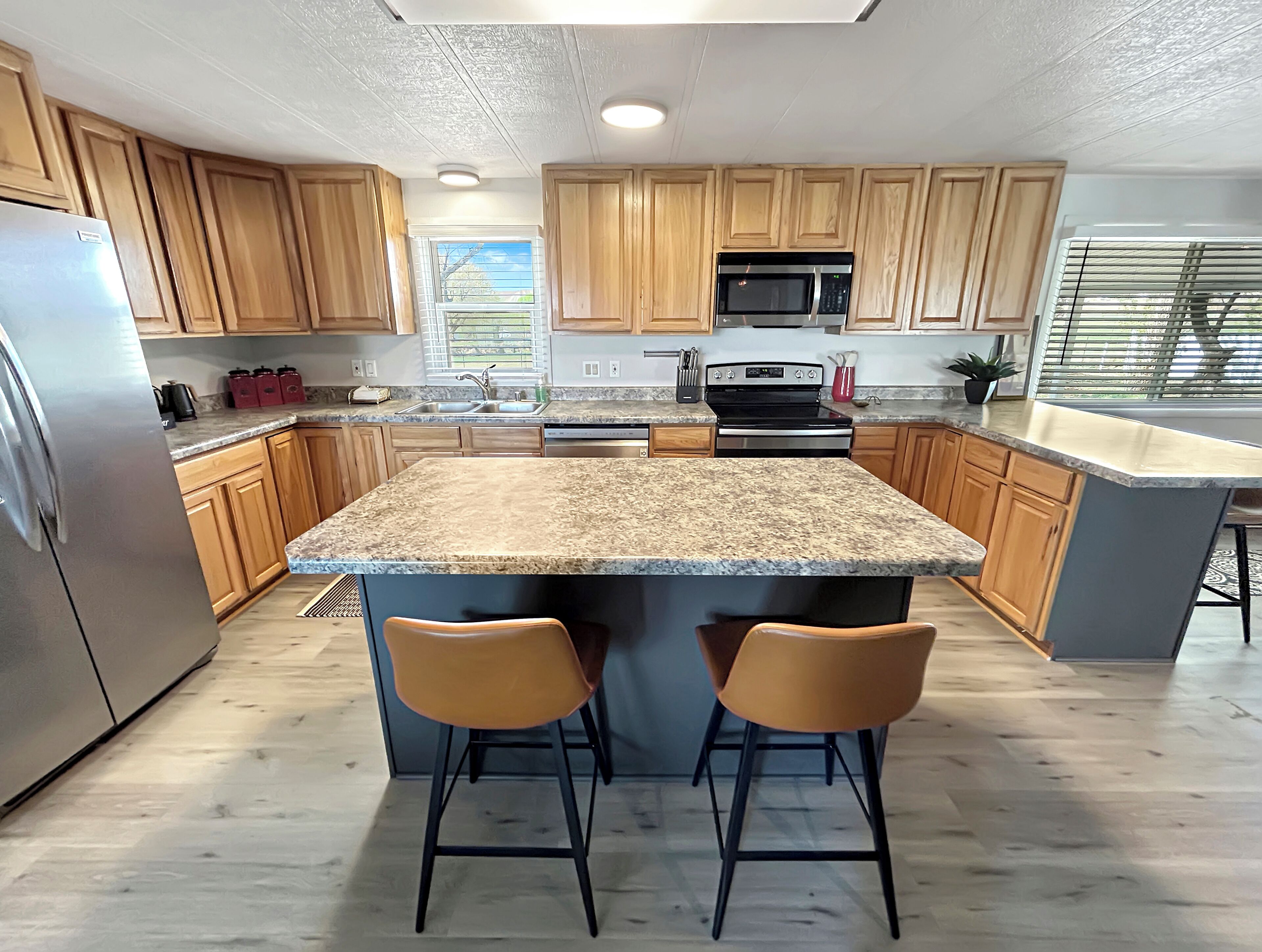A modern kitchen with wooden cabinets, granite countertops, a stainless steel refrigerator, a microwave, and a stove. There are two bar stools at the island.