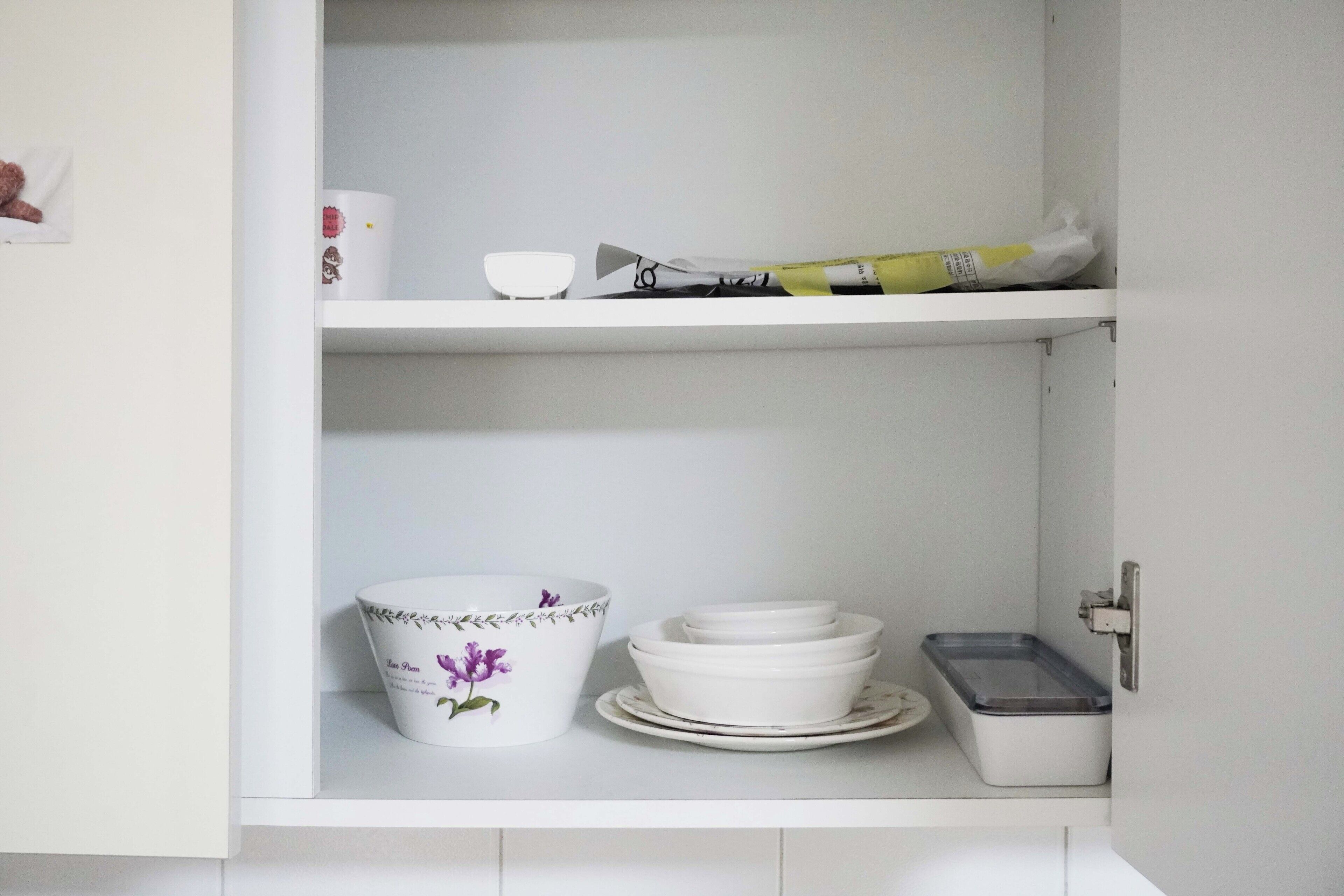 Un meuble de cuisine avec des étagères blanches, contenant un bol avec des fleurs violettes, des bols blancs empilés, une assiette blanche et un récipient blanc avec un couvercle.