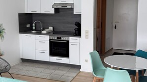 Une cuisine moderne avec des meubles blancs, un plan de travail noir et un four encastré. On y trouve également un coin repas avec une table ronde et des chaises de couleur turquoise.