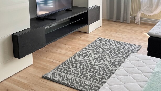 Une chambre moderne avec un téléviseur à écran plat posé sur un meuble noir, une grande fenêtre ornée de rideaux légers et un tapis à motifs sur le plancher en bois.