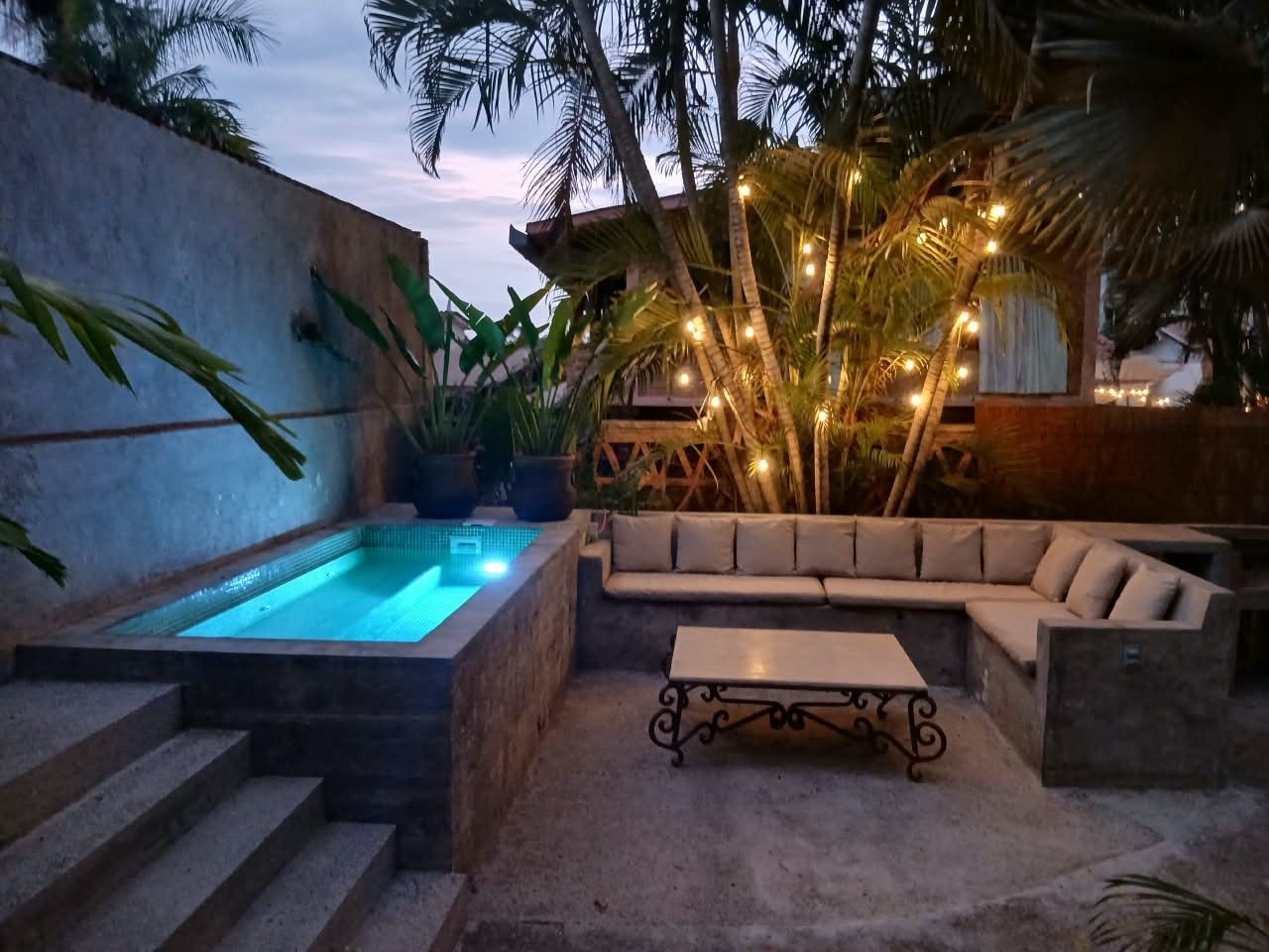 Un espace piscine avec un jacuzzi, un coin salon comprenant un canapé et une table, le tout entouré de plantes et de palmiers.