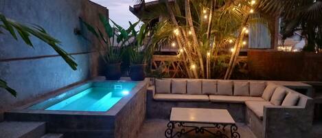 A pool area with a hot tub, a seating area with a sofa and a table, surrounded by plants and palm trees.