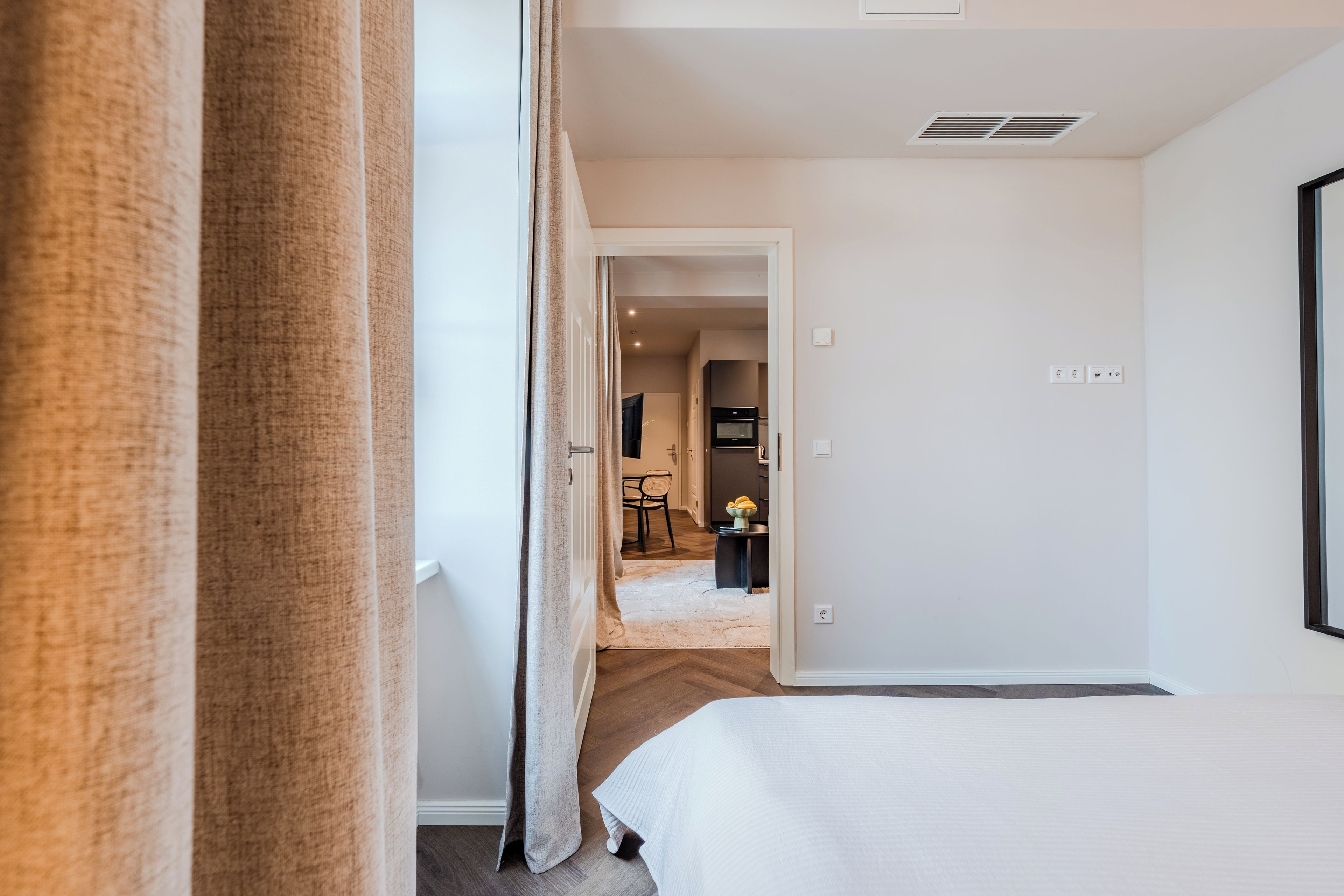 Una habitación de hotel con una cama, una puerta que da a un comedor y una cocina visible al fondo.