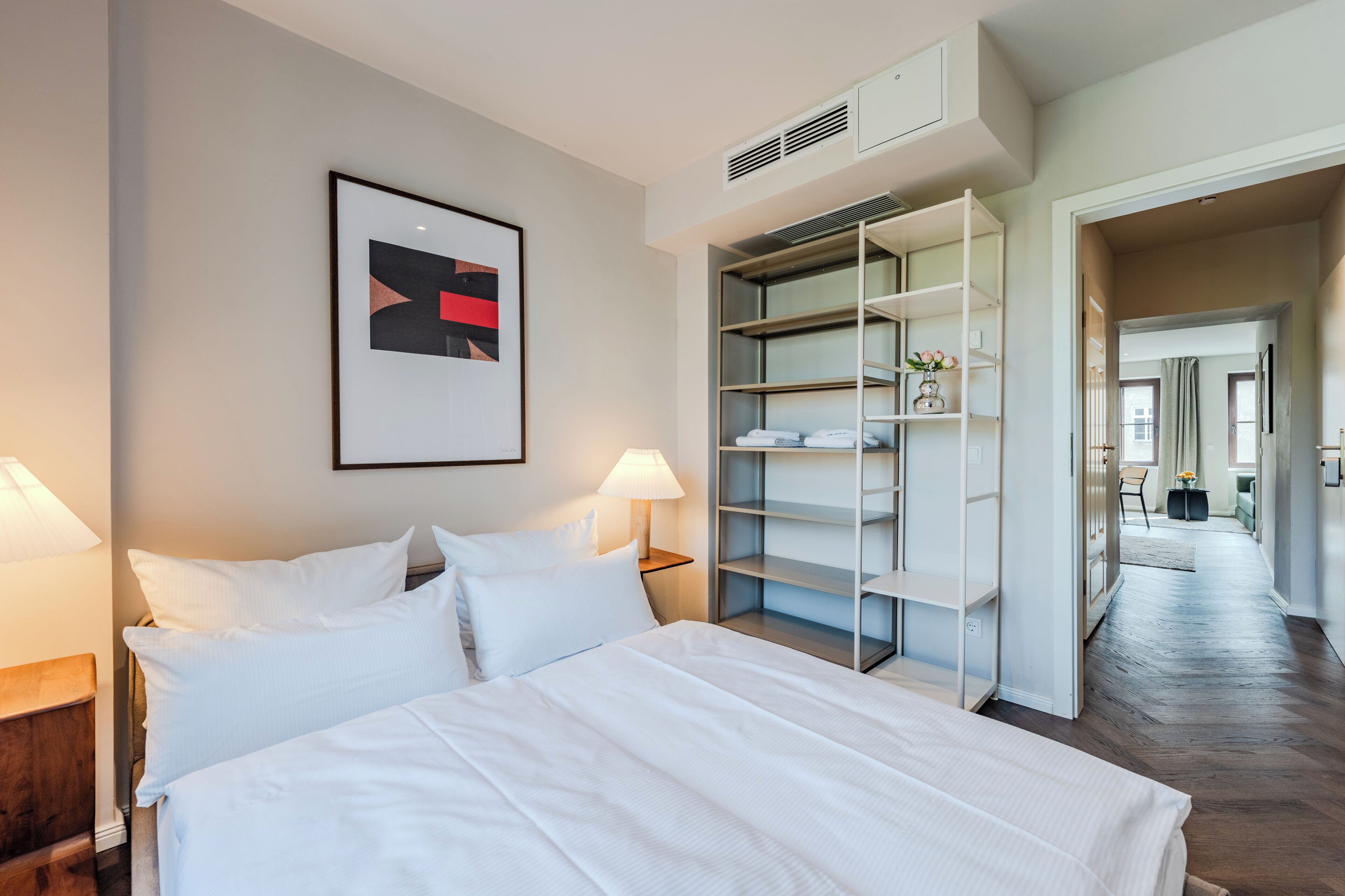 Ein Hotelzimmer mit einem Bett, einem Nachttisch, einer Lampe, einem gerahmten Bild an der Wand und einem begehbaren Kleiderschrank mit Regalen.
