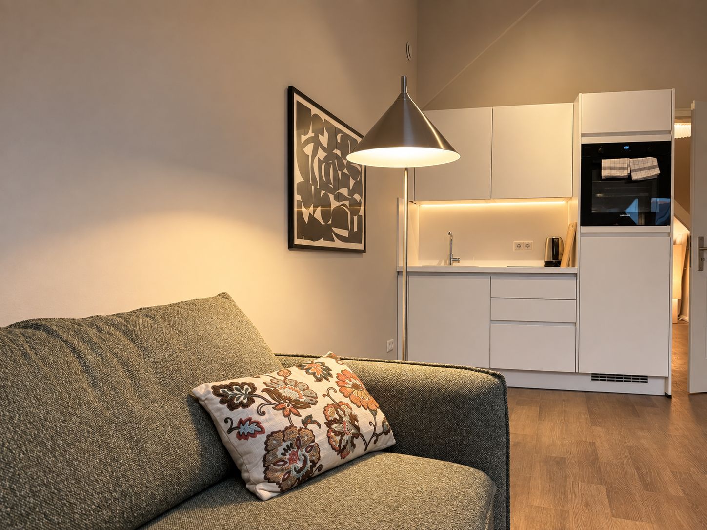 Una sala de estar moderna con un sofá y un cojín decorativo, una lámpara colgante, una zona de cocina con armarios blancos y un horno negro.