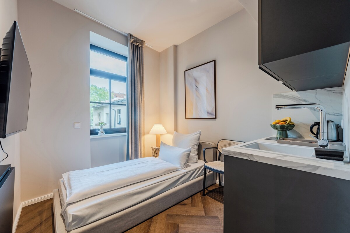 Ein kleines Hotelzimmer mit Bett, einem Schreibtisch mit Lampe, einem Fernseher und einem Fenster mit Vorhängen.