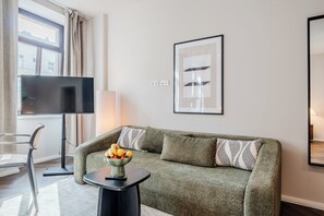 Ein modernes Wohnzimmer mit einem grünen Sofa, einem Beistelltisch mit Obst, einem Flachbildfernseher und einem gerahmten abstrakten Kunstwerk an der Wand.