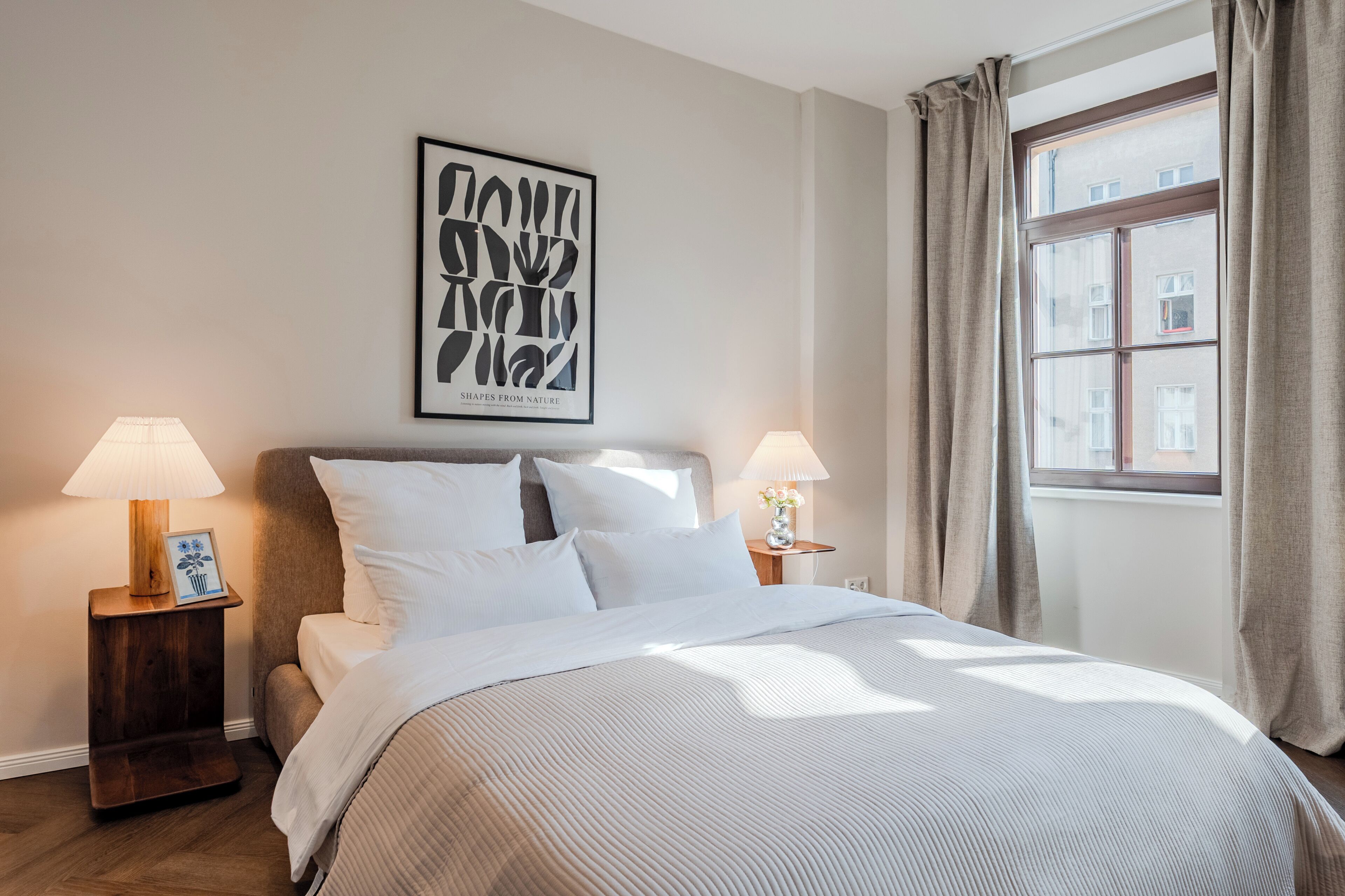 A bedroom with a bed, two bedside tables with lamps, a framed artwork on the wall, and a window with curtains.